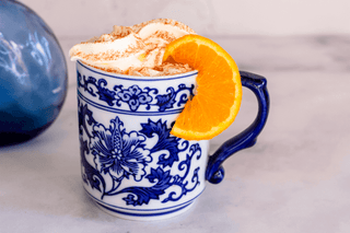 White ceramic cup adorned with blue flower designs. The cup is filled with a drink topped with latte foam and sprinkled with cinnamon.  There is an orange slice garnish on the side of the mug.