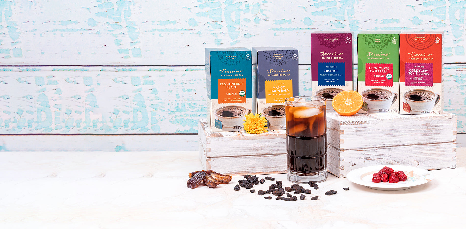 Tall glass with dark iced beverage. Bags and boxes of teeccino flavors, mango lemon balm, Cordyceps, Chocolate raspberry. Ingredients scattered tastefully in front