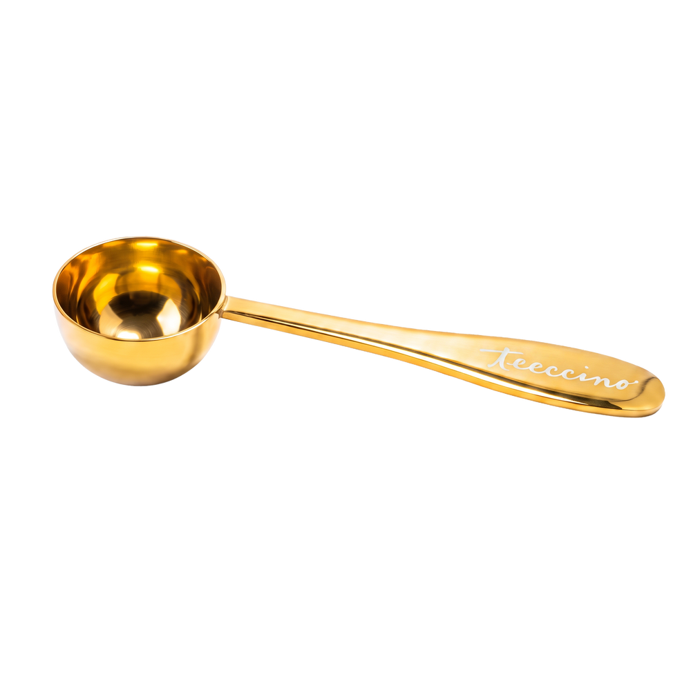 A gold-finish metal measuring scoop with a mirror-polished shine is shown on a transparent background. The scoop features a round bowl and a long, tapered handle engraved with the Teeccino script logo. Designed for portioning herbal coffee or tea, this gold scoop adds a bright, elegant touch to the brewing routine while providing a practical, reusable measuring tool.