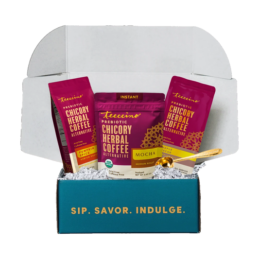 Open teal Teeccino gift box printed “Sip. Savor. Indulge.” holding a Pumpkin Spice Chicory Herbal Coffee bag, an Instant Mocha Chicory Herbal Coffee pouch, a Maple Pecan Chicory Herbal Coffee bag and a gold Teeccino scoop on white crinkle filler, on a transparent background.