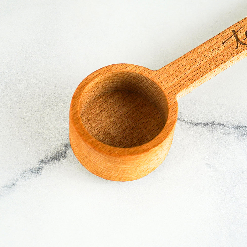 Top-down view of a round wooden measuring scoop with a long handle, resting on a white marble surface with a grey vein. The scoop is carved from light-colored wood with a visible grain. Part of a dark cursive logo is visible on the handle's right side. The lighting is bright and even, highlighting the interior texture of the scoop.