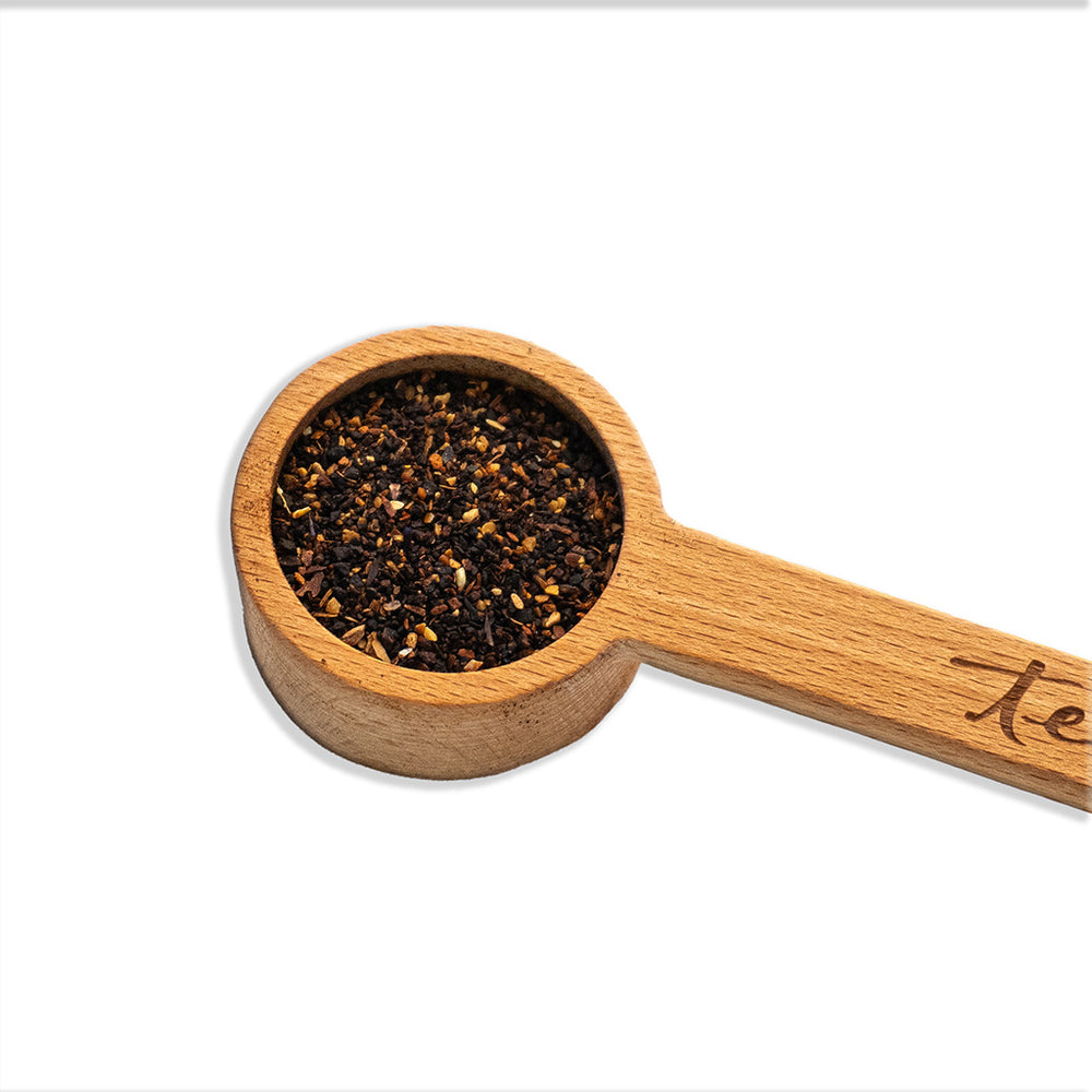 Top-down view of a round wooden measuring scoop with a long handle, resting on a white marble surface with a grey vein. The scoop is carved from light-colored wood with a visible grain. Part of a dark cursive logo is visible on the handle's right side. The lighting is bright and even, highlighting the interior texture of the scoop.