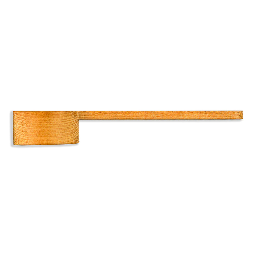  wooden measuring scoop with a long handle, resting on a white marble surface with a grey vein. The scoop is carved from light-colored wood with a visible grain. Part of a dark cursive logo is visible on the handle's right side. The lighting is bright and even, highlighting the interior texture of the scoop.