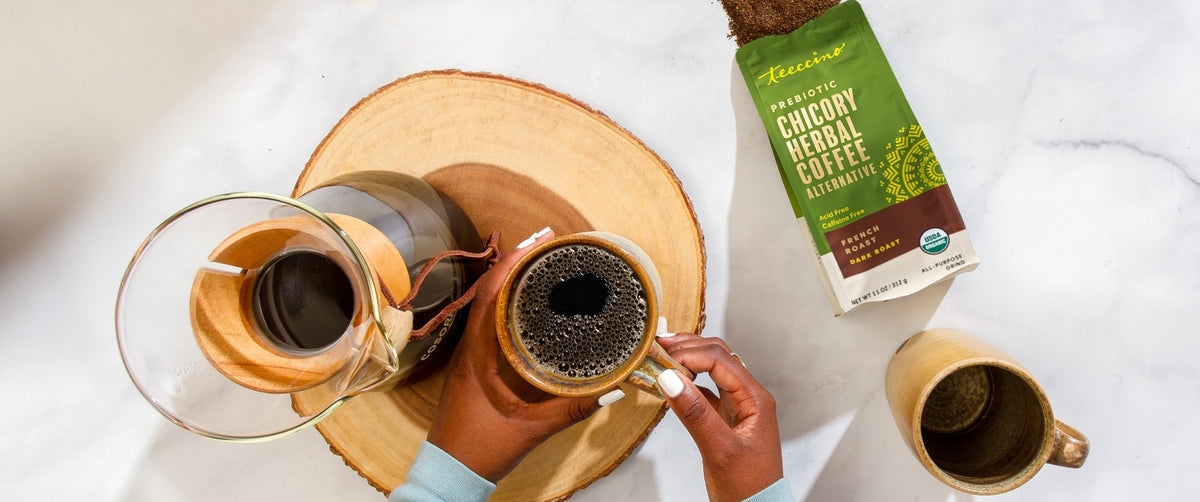 A bag of Teeccino French Roast Chicory Herbal Coffee beside a glass pour-over carafe and a mug of dark brewed coffee on a wood slice.